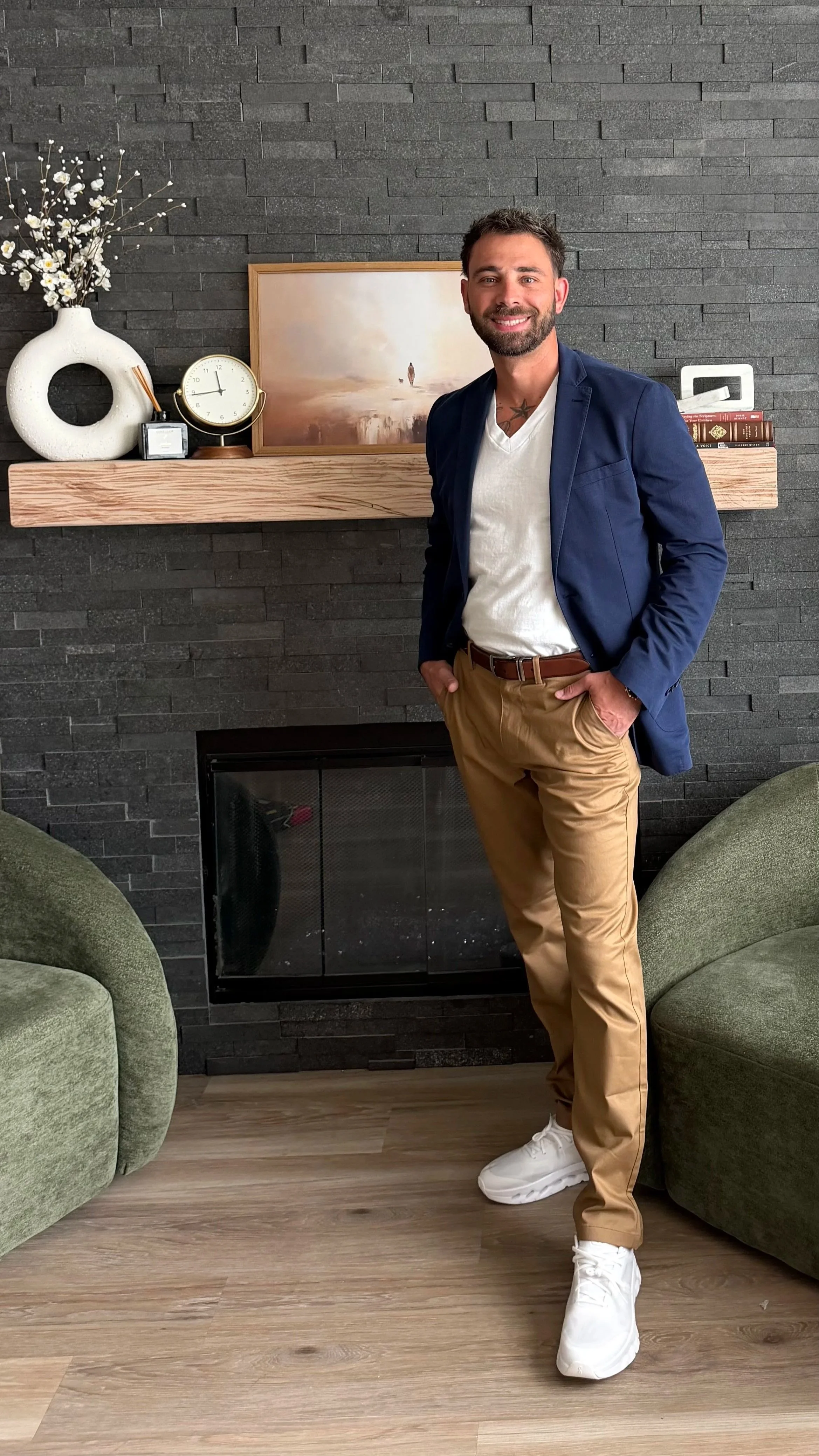 A man smiling and standing indoors in front of a dark gray brick wall. He is wearing a navy blazer, white t-shirt, khaki pants, and white sneakers. There is a wooden mantel above a black fireplace, decorated with a white vase with flowers, a round clock, and a couple of books. To the sides of the man, there are green armchairs.