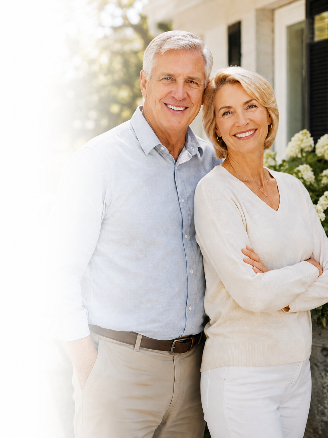 A happy older couple standing outside in front of a house with white flowers, smiling at the camera.