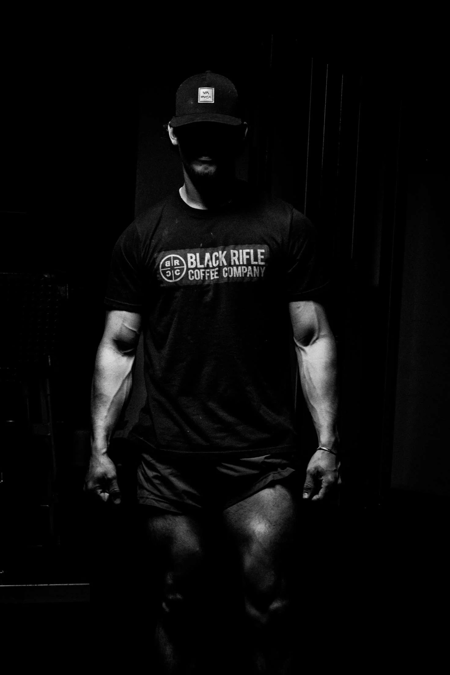 Black and white photo of a muscular man wearing a cap and t-shirt with the logo 'Black Rifle Coffee Company', sitting with fists clenched, in a dark room.
