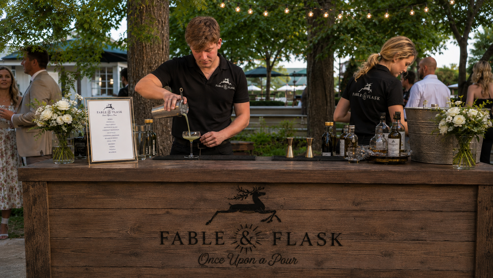 Two bartenders at an outdoor event behind a wooden bar with bottles and floral arrangements, with guests in the background.