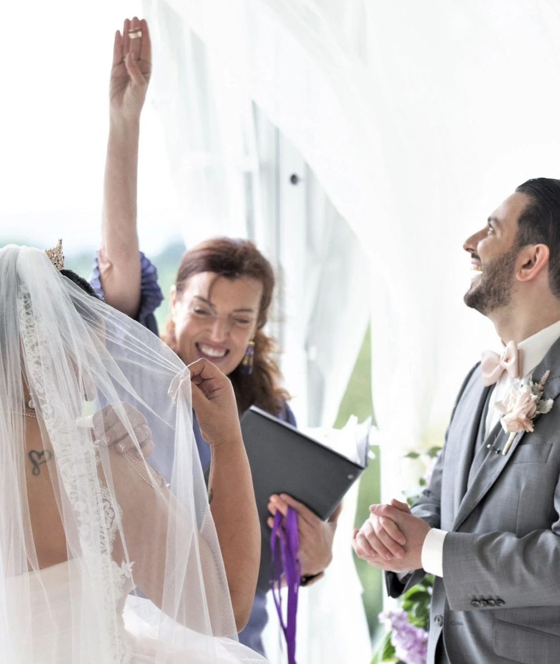 A wedding ceremony with a bride, groom, and officiant indoors; the bride is in a wedding dress with a veil, the groom is in a gray suit with a pink bow tie, and the officiant is holding a book and smiling.
