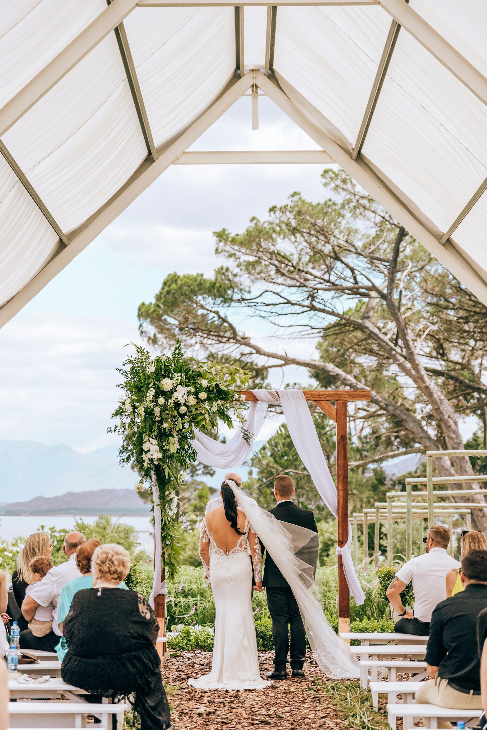 A bride and groom exchange vows under an outdoor wedding arch decorated with white drapery and floral arrangements, with guests seated on either side and scenic trees and mountains in the background.