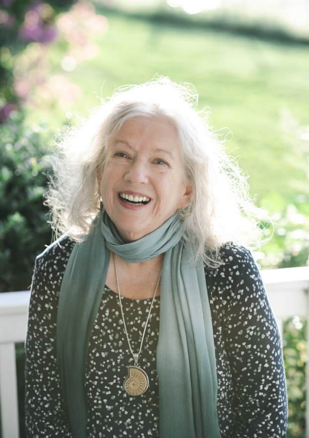 A smiling elderly woman with white hair, wearing a black and white patterned top, a gray scarf, and a necklace with a large shell pendant, standing outdoors with green foliage in the background.