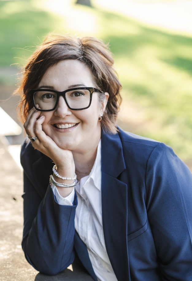 A woman with short brown hair, glasses, and a friendly smile, dressed in a dark blue blazer and white shirt, sitting outdoors with a blurred green background.