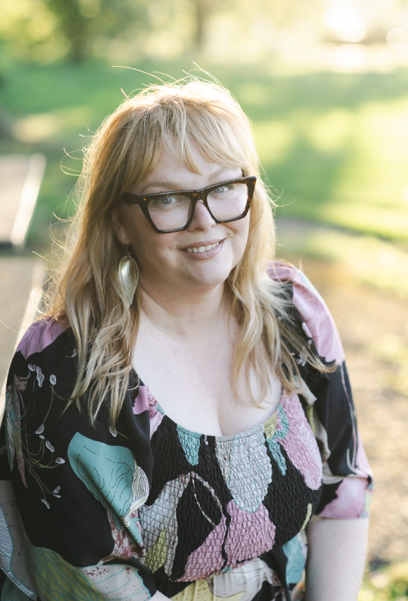 A woman with blonde hair, glasses, and earrings smiling outdoors with green trees and sunlight in the background.