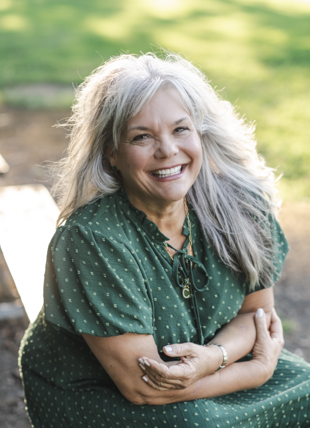Smiling woman with gray hair sitting outdoors on a sunny day, wearing a green dress with small yellow polka dots.