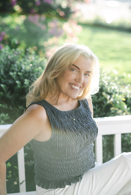 A smiling middle-aged woman with blonde hair sitting outdoors on a white bench, wearing a sleeveless dark top and white pants, with a green garden and pink flowers in the background.