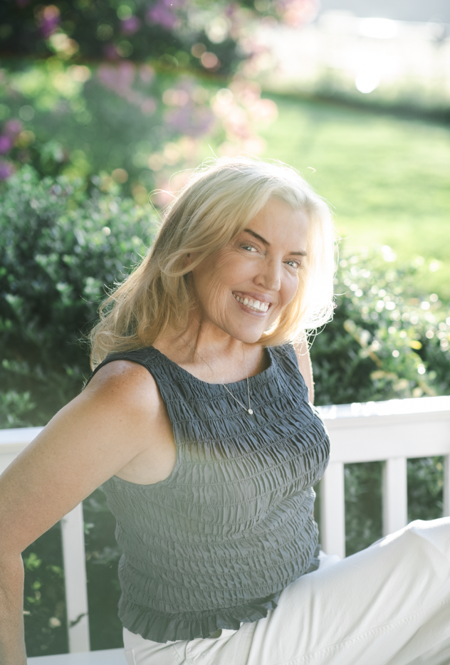 A smiling blonde woman sitting outside on a porch with a white railing, greenery, and pink flowers in the background.