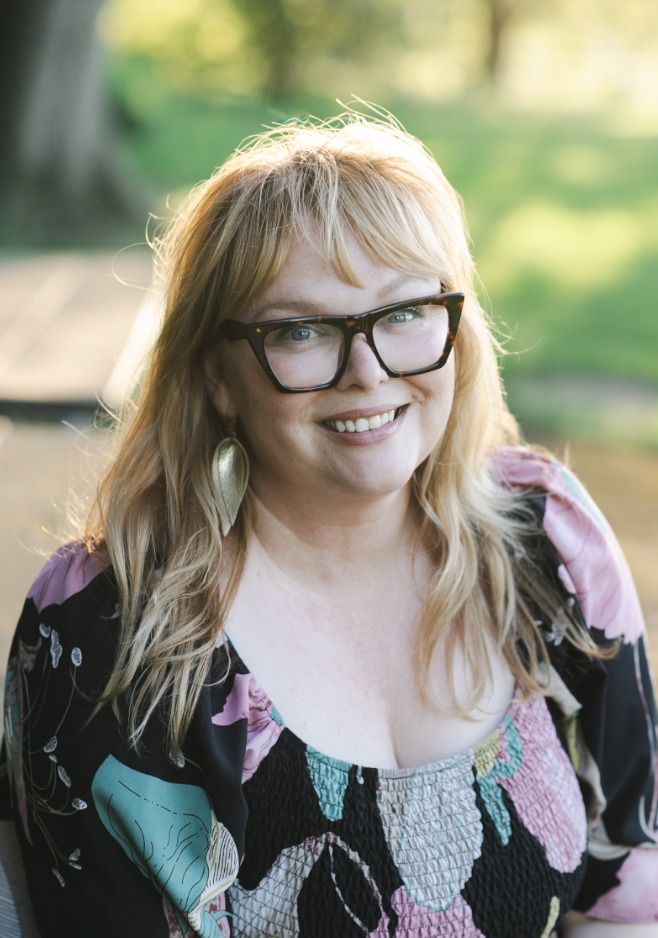A smiling woman with glasses and blonde hair outdoors on a sunny day.
