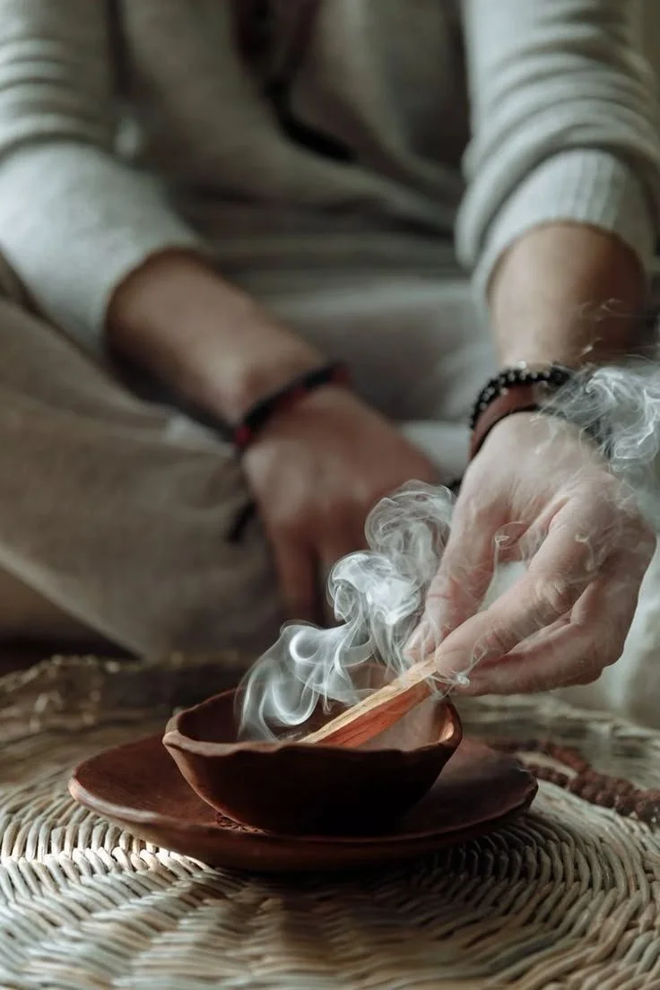 Close-up of a person holding a burning incense stick over a small burning bowl, with wisps of smoke rising from it.