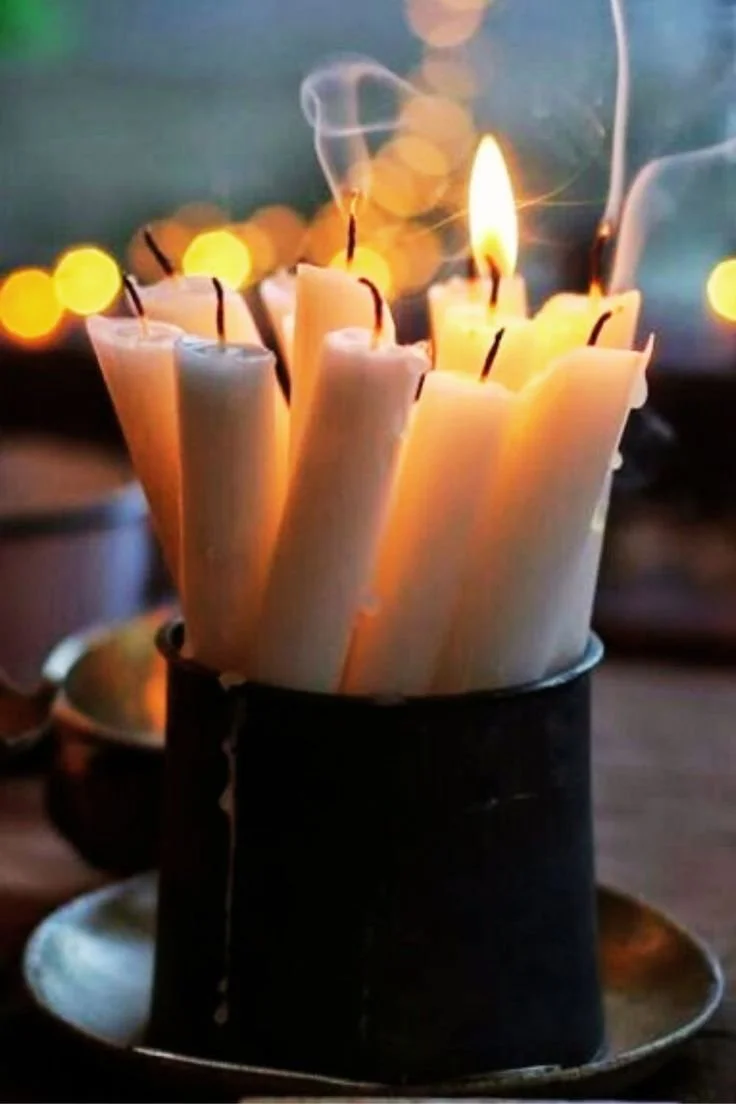 Group of candles with one lit on a dark surface, surrounded by blurred warm lights in the background.