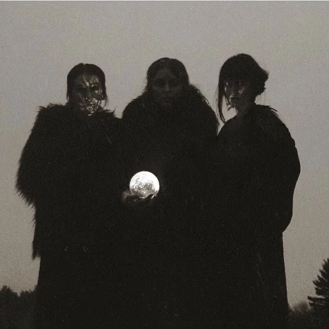 Three people standing outdoors at night, with the moon visible behind them, dressed in dark clothing.