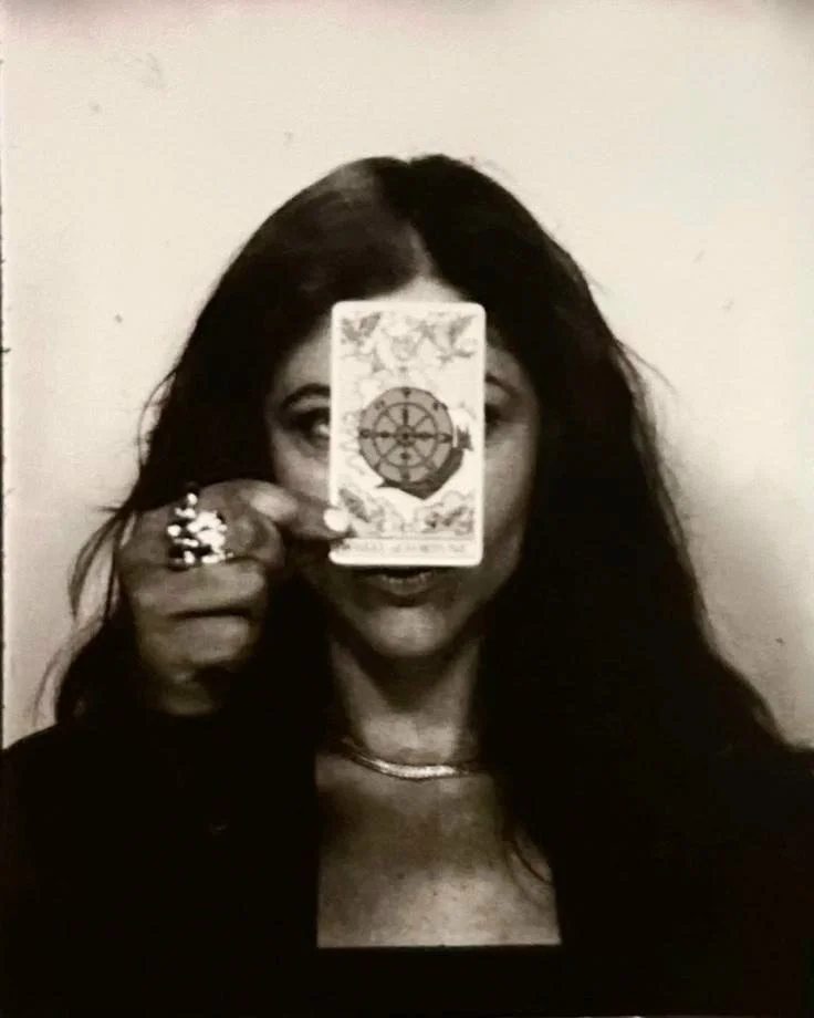 Black and white photo of a woman with long dark hair holding a tarot card in front of her face, partially obscuring it.
