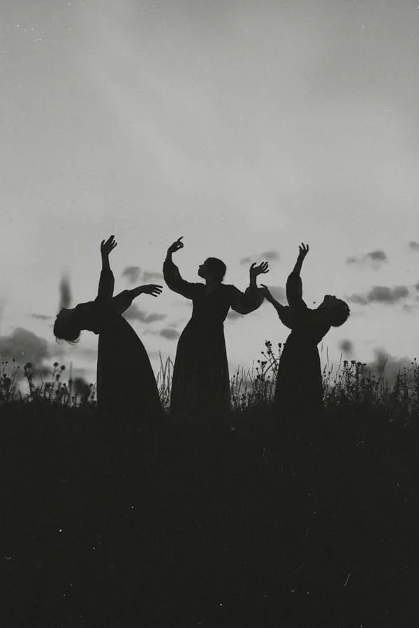 Silhouettes of three women standing in a field at sunset, with their arms raised and bent in artistic poses.