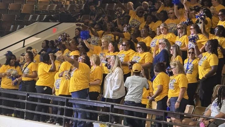 Group of people wearing yellow shirts gathered in a stadium, some holding blue and gold pom-poms, appearing to cheer or celebrate.