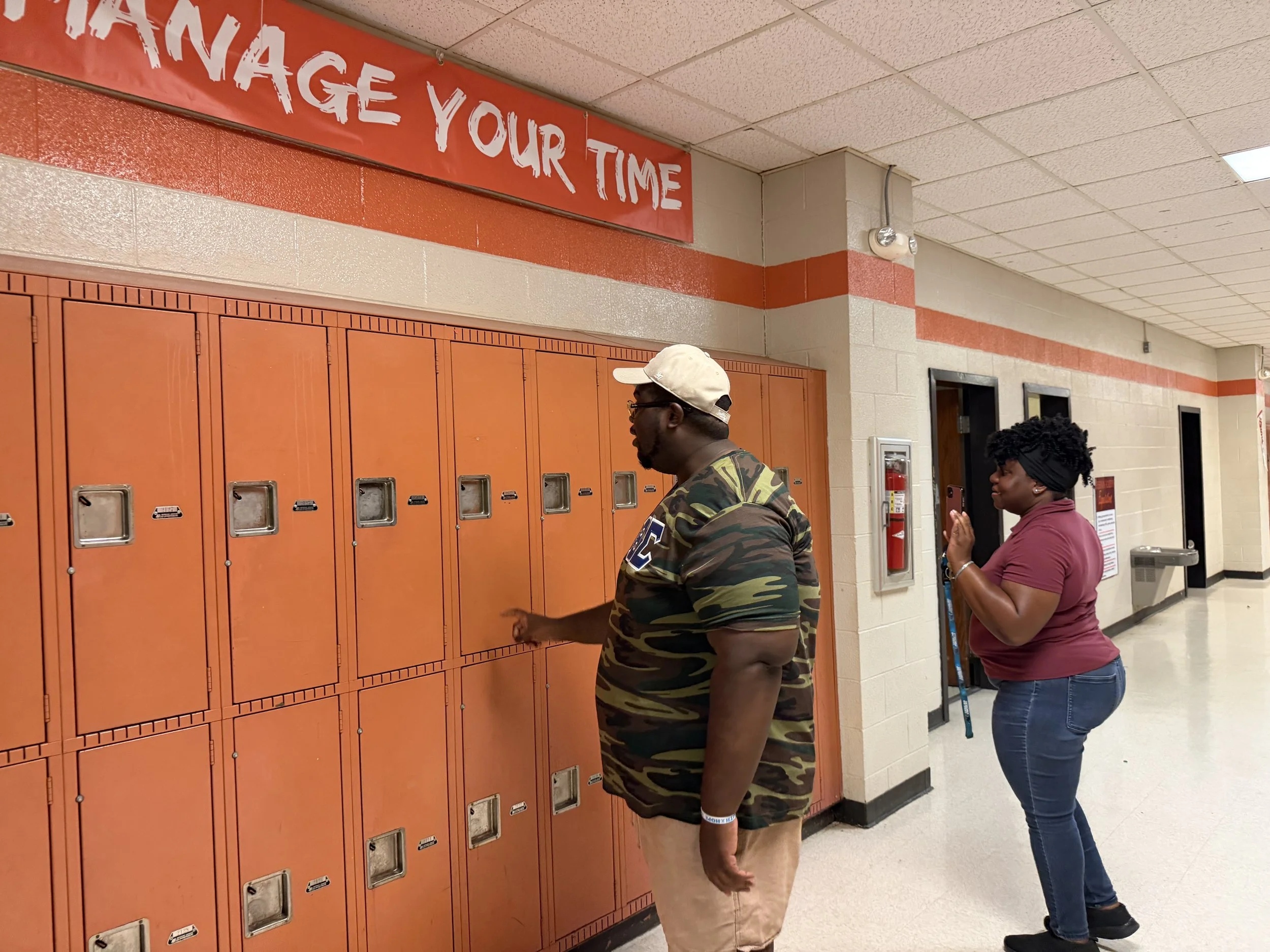 Students at lockers in a school hallway, one male student wearing a camouflage shirt and another female student in a purple top. There is a red banner above the lockers that reads 'Improve Your Time'.