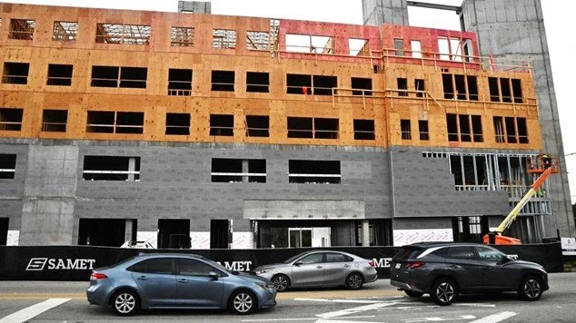 Construction site of a multi-story building with workers and equipment, tall cranes, and parked cars in front.