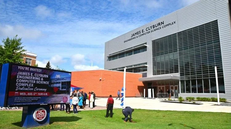 Group of people gather outside James E. Clyburn Engineering & Computer Science Complex building with digital invitation board displaying an event announcement.