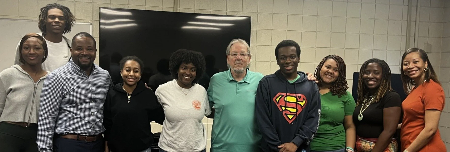 Group of eleven diverse people, including young adults and an older man, standing together in an indoor room with a large black screen behind them, posing for a photo, smiling.