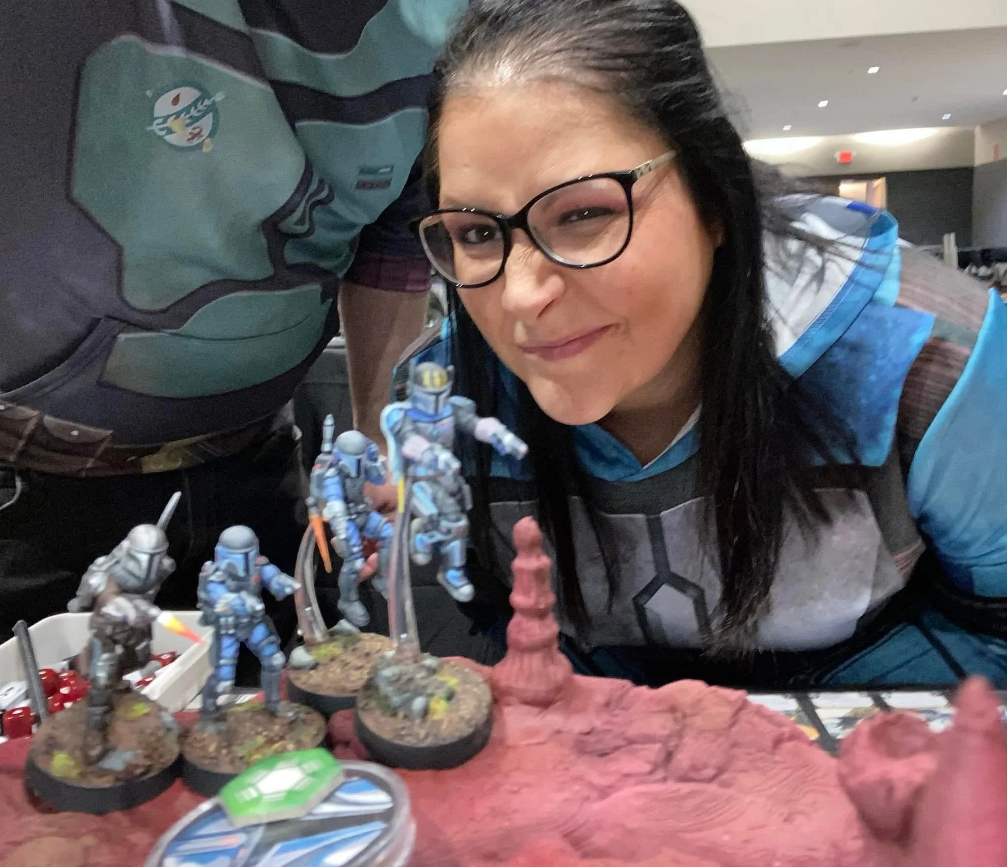 A woman with glasses and dark hair, smiling and resting her head on her hand, is at a table filled with small toy soldiers, tanks, and figurines, likely modeling or gaming.