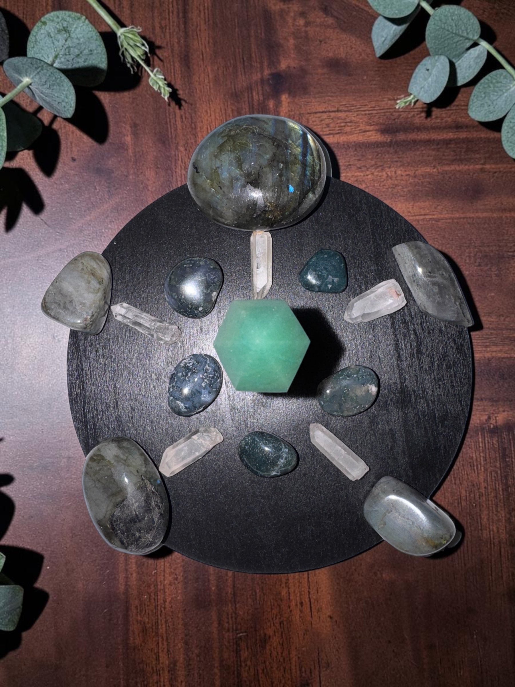 A circular black table with various polished stones and crystals, including a large round stone, a green hexagonal crystal, and several elongated quartz points, on a wooden surface, with green eucalyptus leaves around the edges.