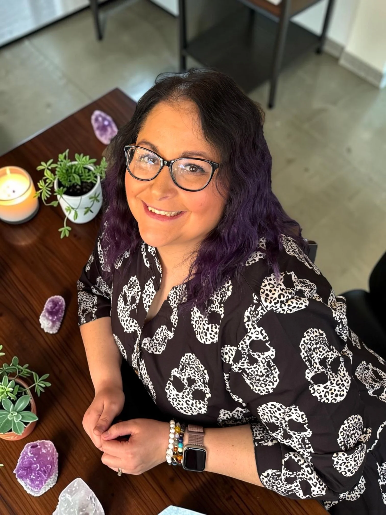 A woman with black, wavy hair and glasses, smiling and wearing a patterned black and white blouse, is sitting at a wooden table with various crystals and small potted plants, in an indoor setting with black chairs and a light-colored floor.