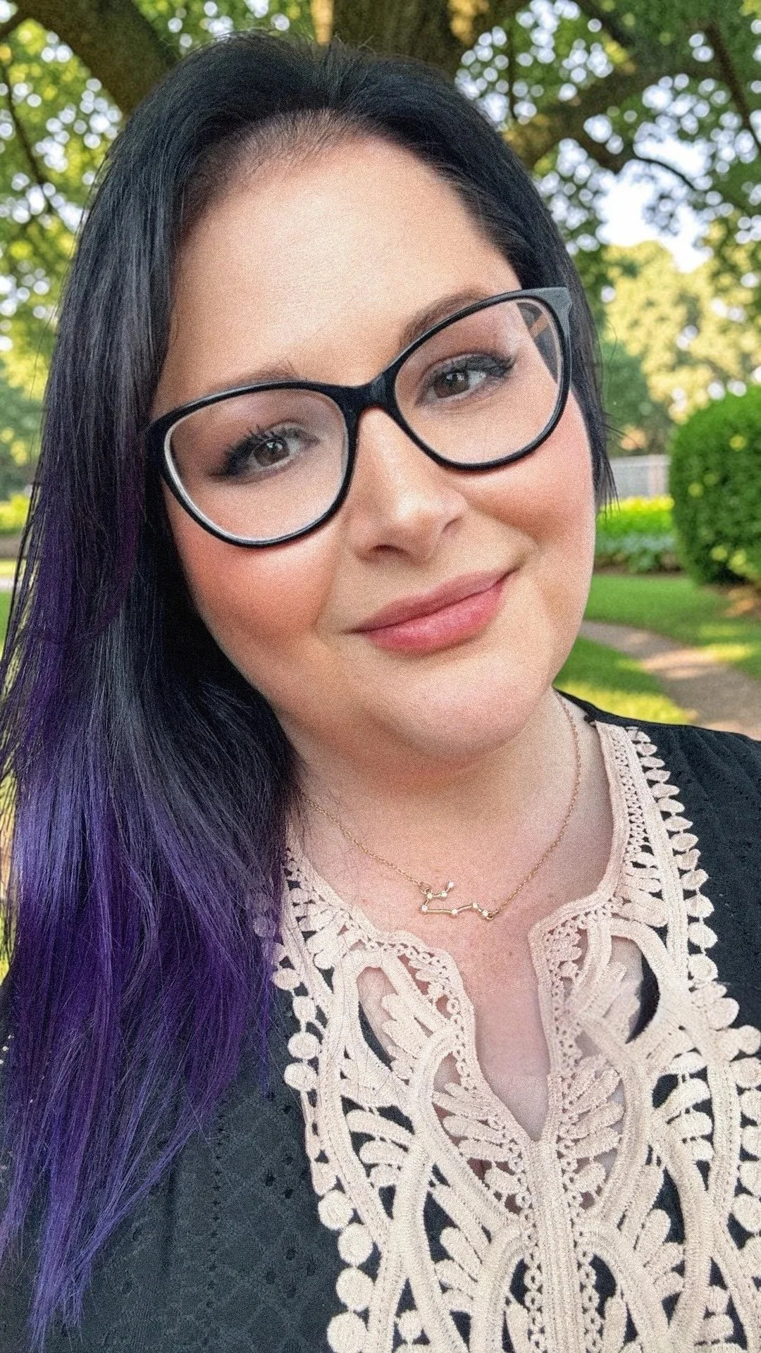 A woman with black hair and purple streaks, wearing large black glasses, a necklace, and an intricate beige lace top, standing outdoors in a green park with trees and a pathway in the background.