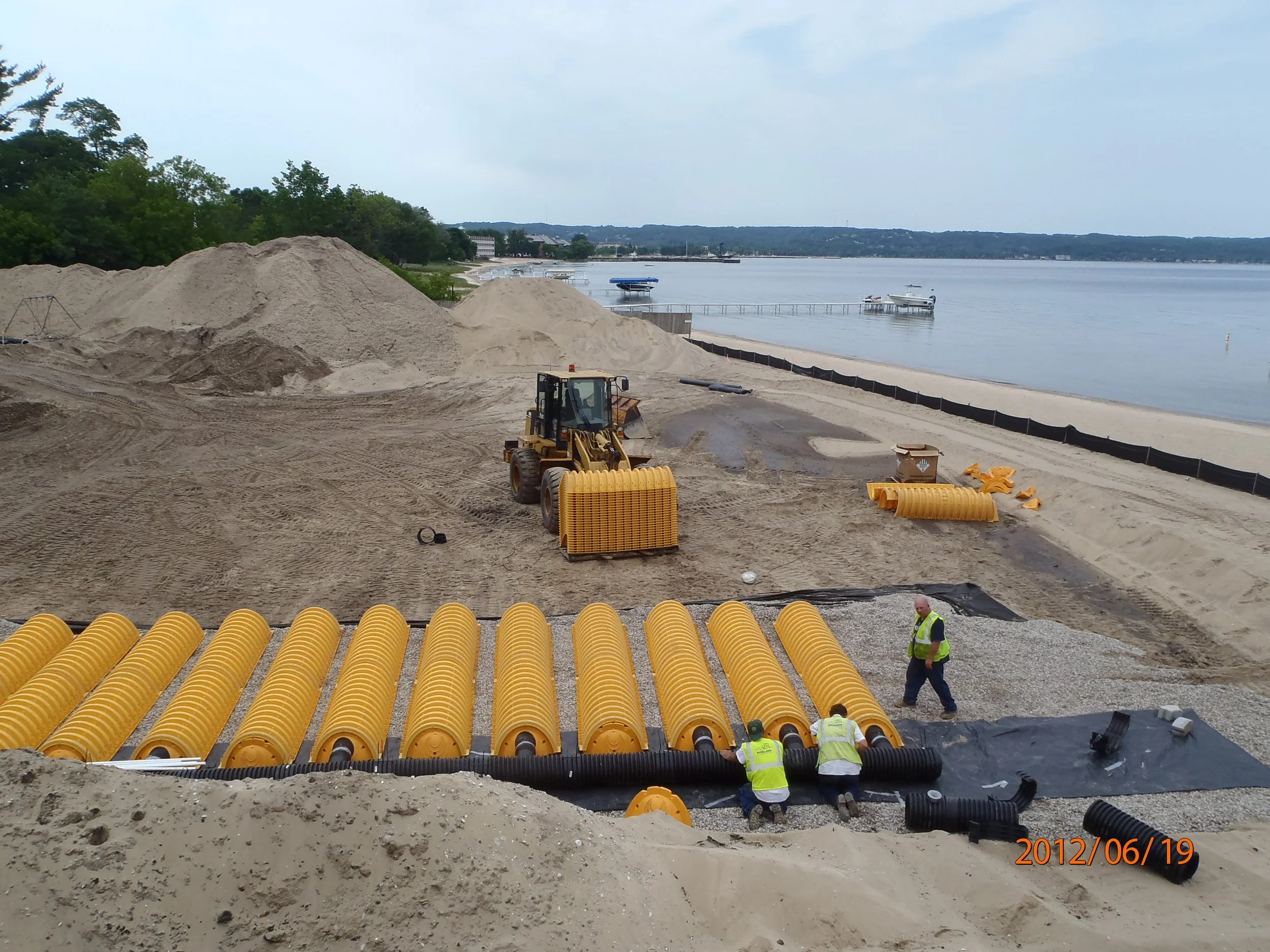 Construction workers installing large yellow drainage pipes along a beach side, with piles of sand, a bulldozer, and a calm water body in the background.