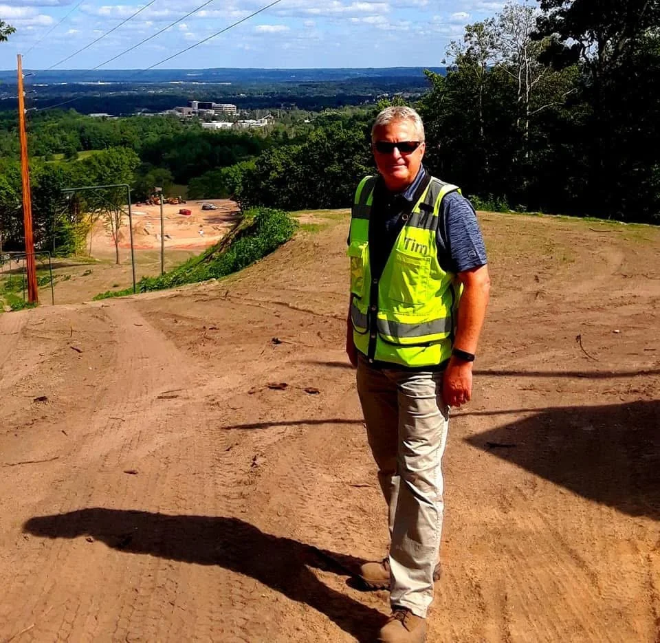 A man wearing sunglasses, a blue shirt, khaki pants, and a yellow safety vest stands outdoors on a dirt construction site with a cityscape in the background.