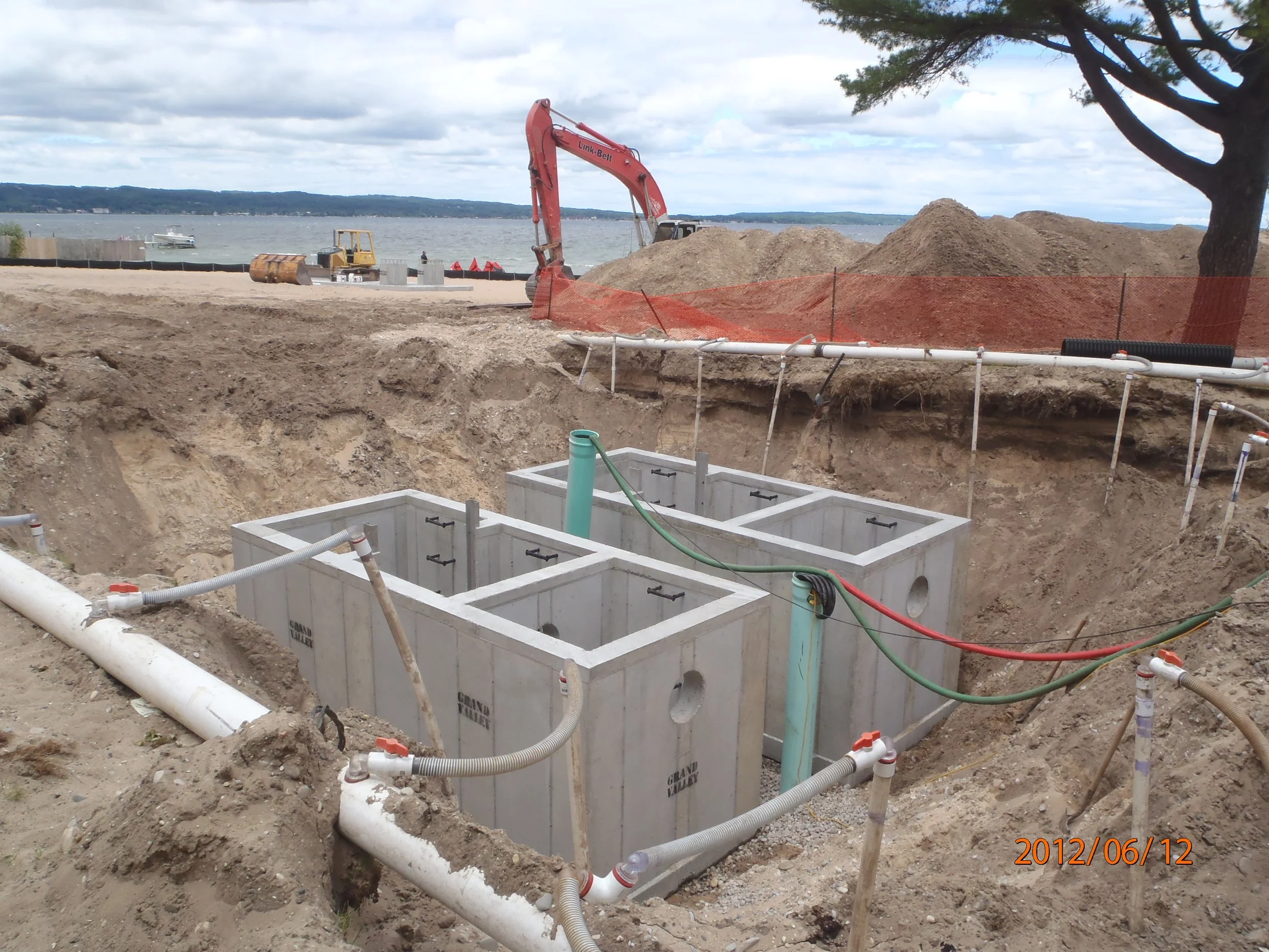 Under construction site near water with excavation, pipes, and concrete foundations, with a tree and piles of dirt in the background.
