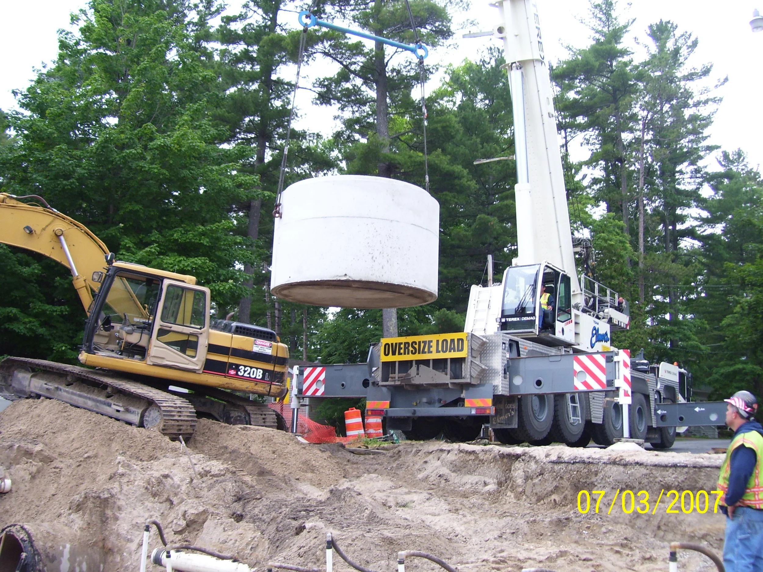 Construction scene with a crane lifting a large white concrete cylinder. There is a yellow excavator on the left and a worker wearing a high-visibility vest and helmet near the bottom right. The area is outdoors with green trees in the background. A caution sign on the crane reads 'Over Size Load', and orange barriers are visible.
