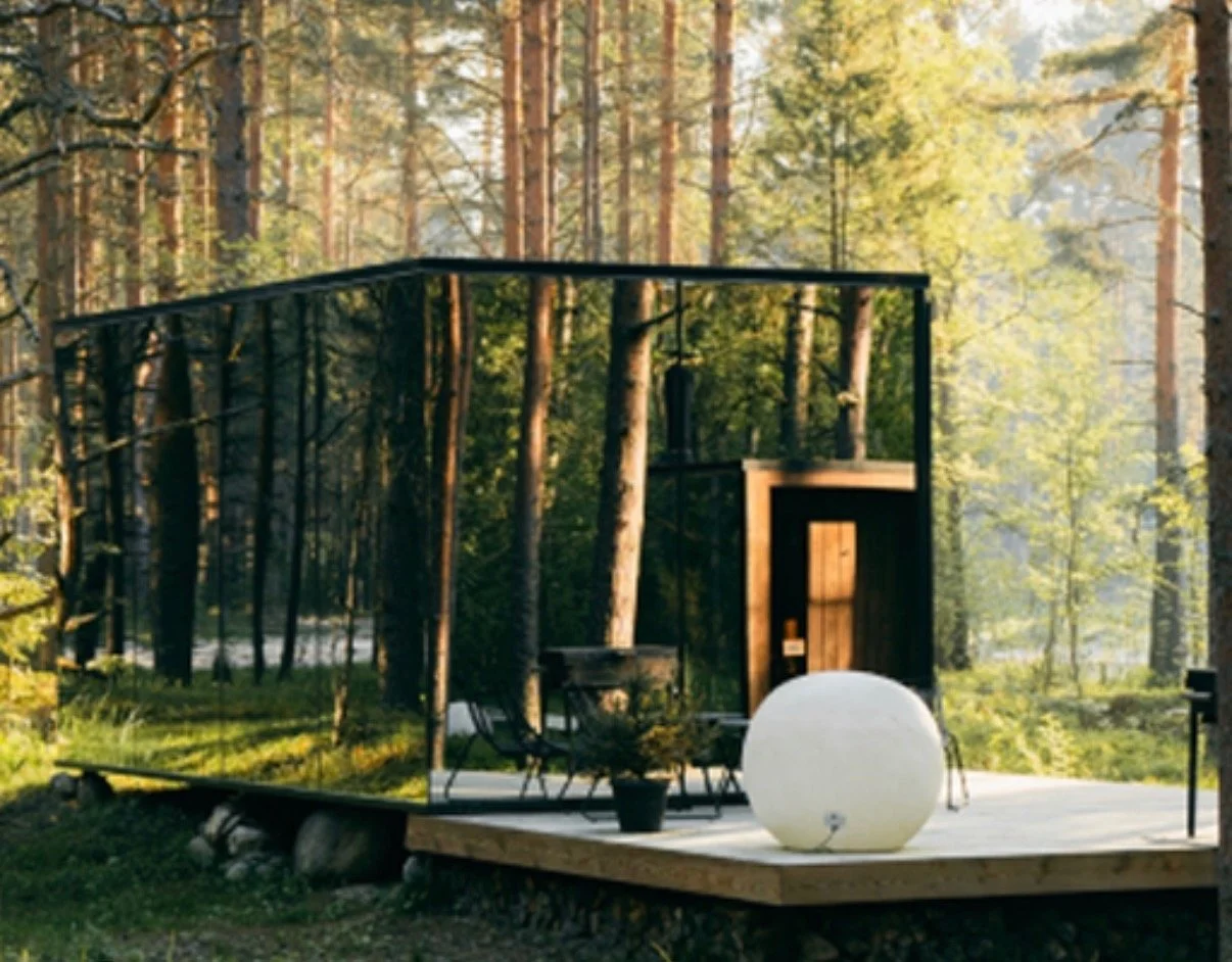 Modern outdoor deck in a forest surrounded by tall trees, featuring a large white sphere, potted plants, outdoor furniture, and a black metal framed glass enclosure.