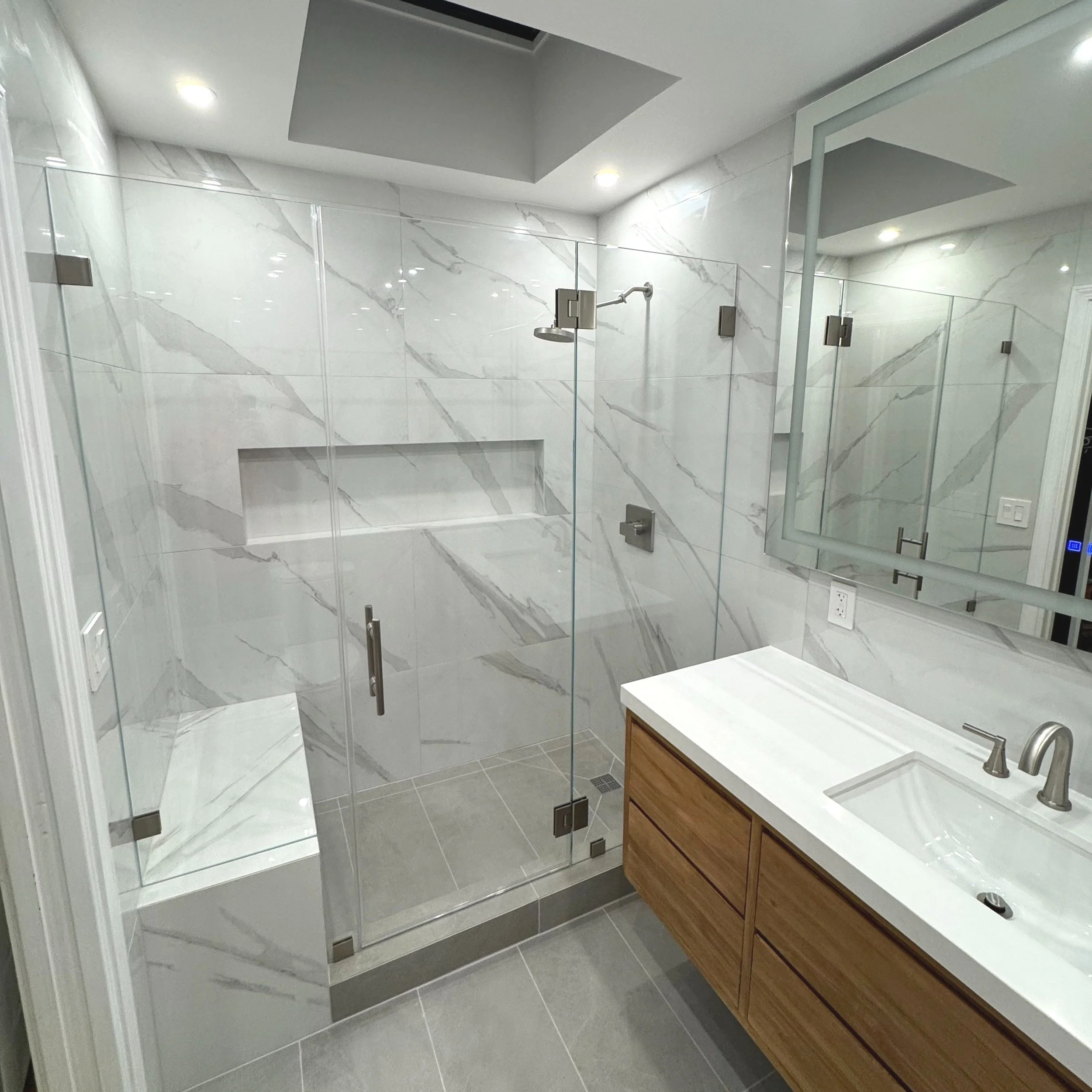 Modern bathroom renovation in San Francisco, glass-enclosed shower featuring marble walls, built-in niche, and gray tile floor. Wooden vanity with a white countertop and a large rectangular mirror. Recessed ceiling lights illuminate the space.