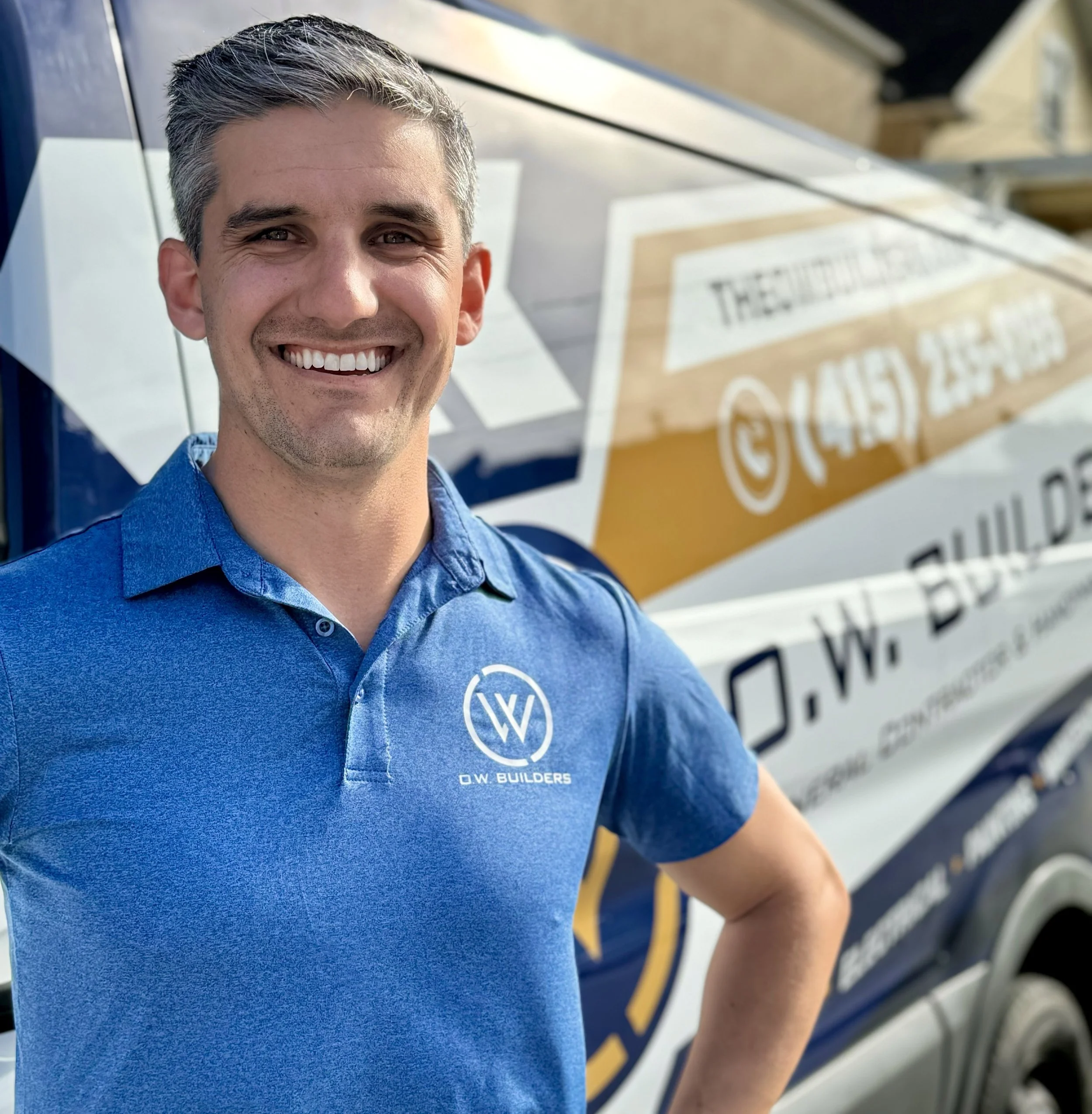 Man smiling in front of a van with branding, wearing a blue polo shirt with the logo 'O.W. BUILDERS'.
