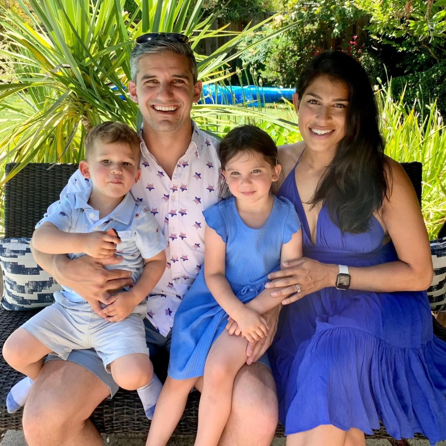 A family of five sitting on a black woven outdoor sofa, smiling at the camera, surrounded by lush green plants and trees in a backyard garden.