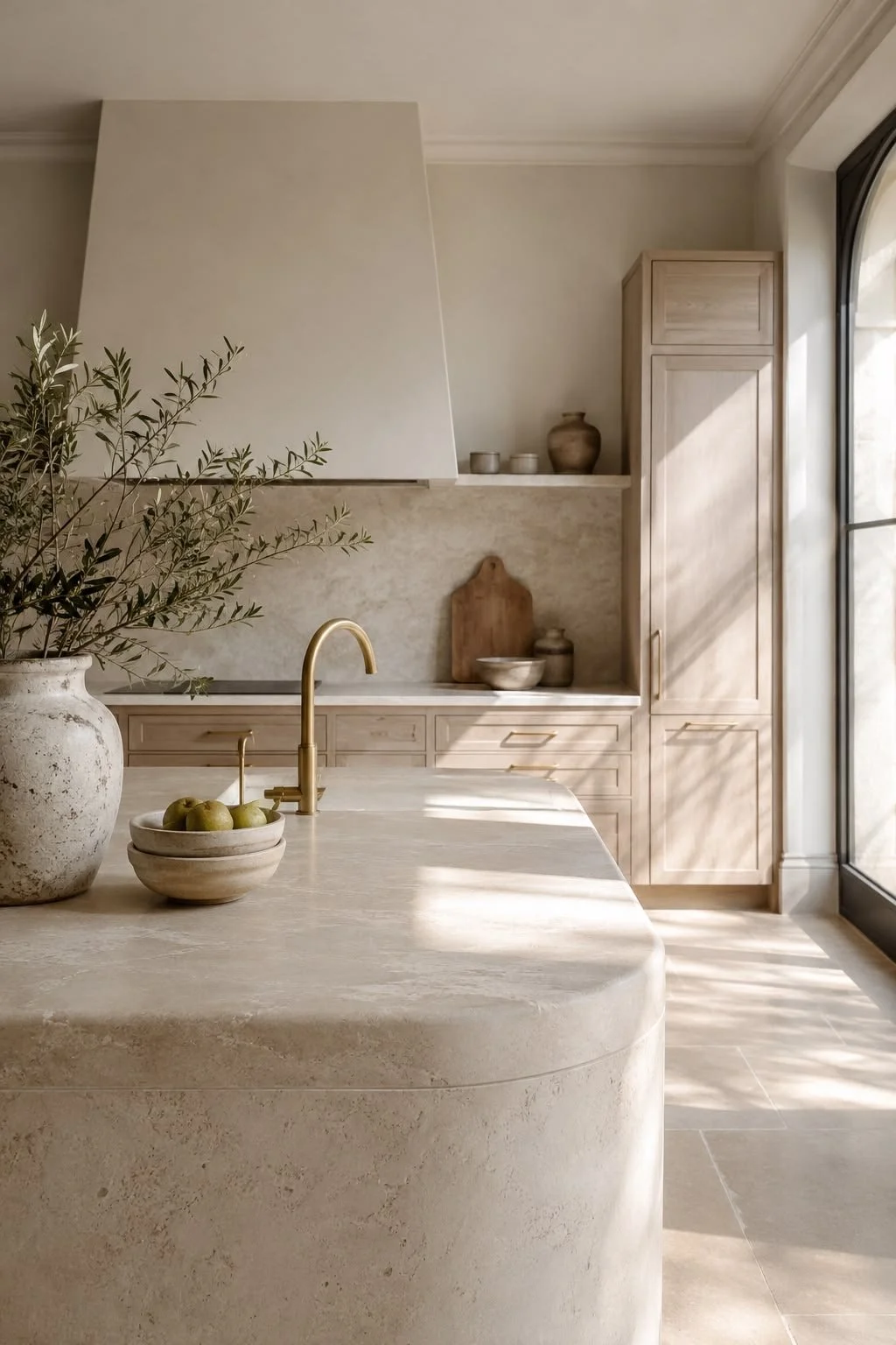 A modern kitchen with beige cabinets, a large window with sunlight shining through, a marble countertop, a gold faucet, a bowl of green apples, decorative pottery, and a large potted olive branch.