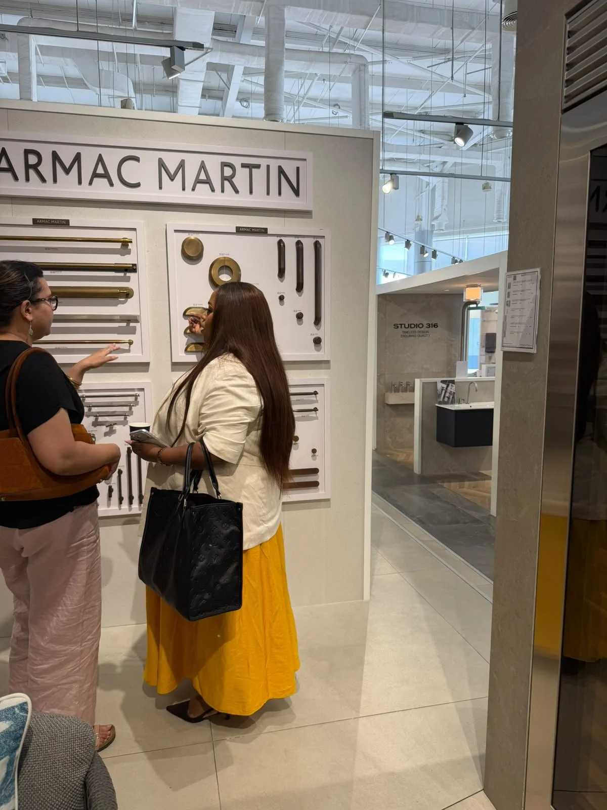 Two women are discussing hardware and fixtures from Armac Martin at a showroom. One is wearing glasses, a black top, and pink pants, holding a brown bag. The other has long brown hair, a white blazer, a yellow skirt, and a black handbag. The display showcases various metallic hardware samples against a white background.