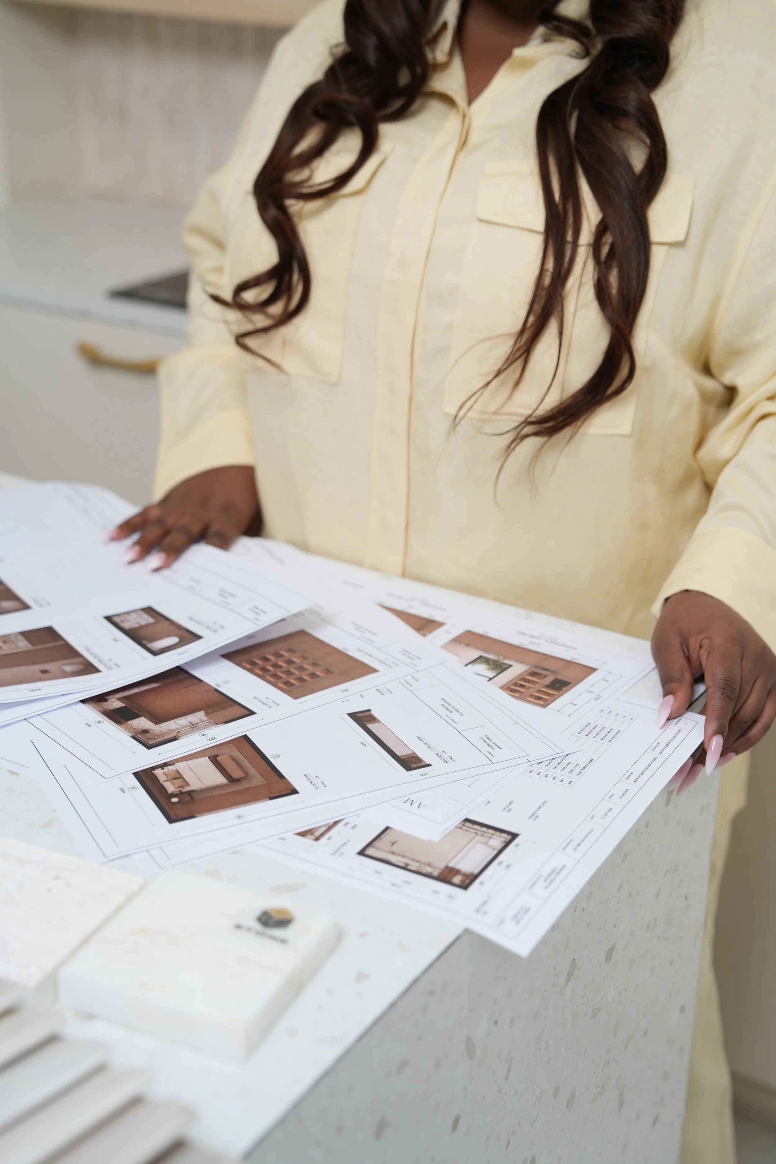 A woman with long curly hair wearing a yellow shirt is reviewing printed interior design plans or catalogs spread out on a white countertop.