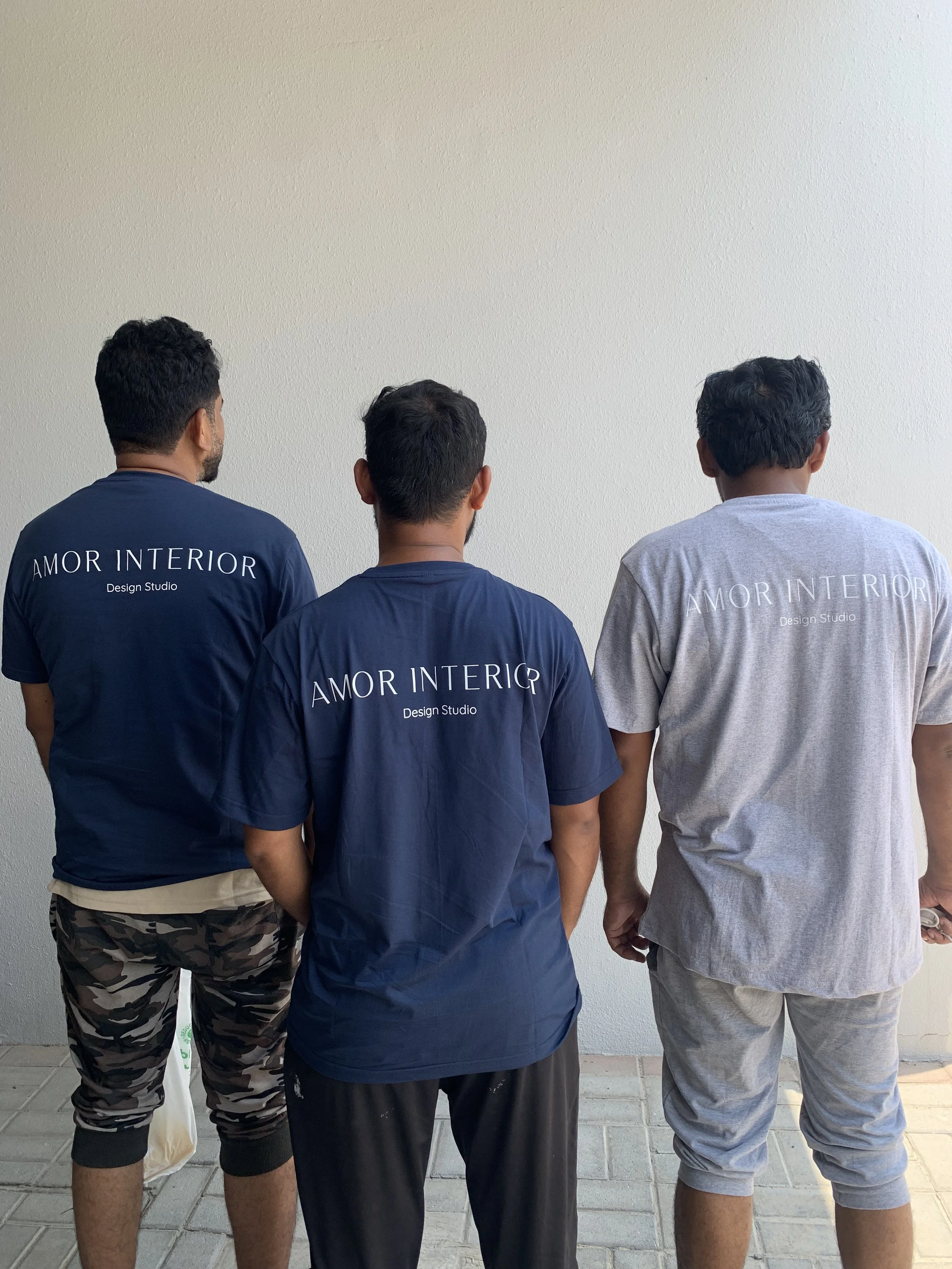 Three men standing outdoors against a neutral wall, viewed from the back, wearing casual clothing with 'Amor Interior Design Studio' printed on their shirts.