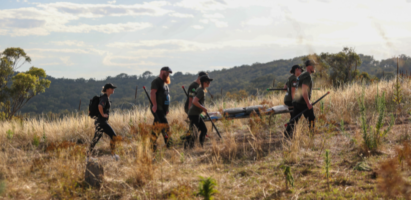 30 Unbroken participants walking through a grassy field, carrying equipment, with hills and trees in the background.