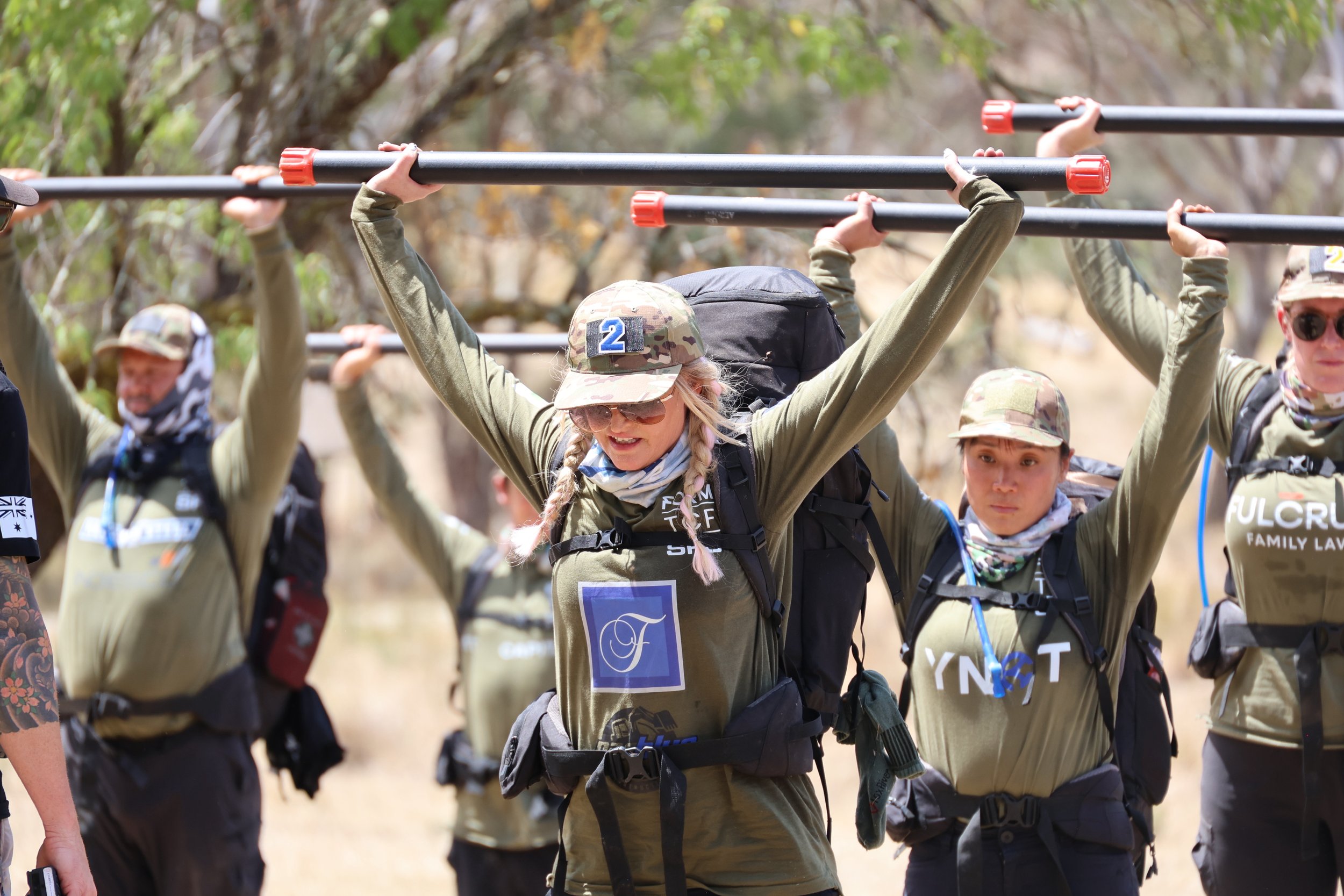30 Unbroken participants in an outdoor activity, carrying bars over their heads with a background of trees.