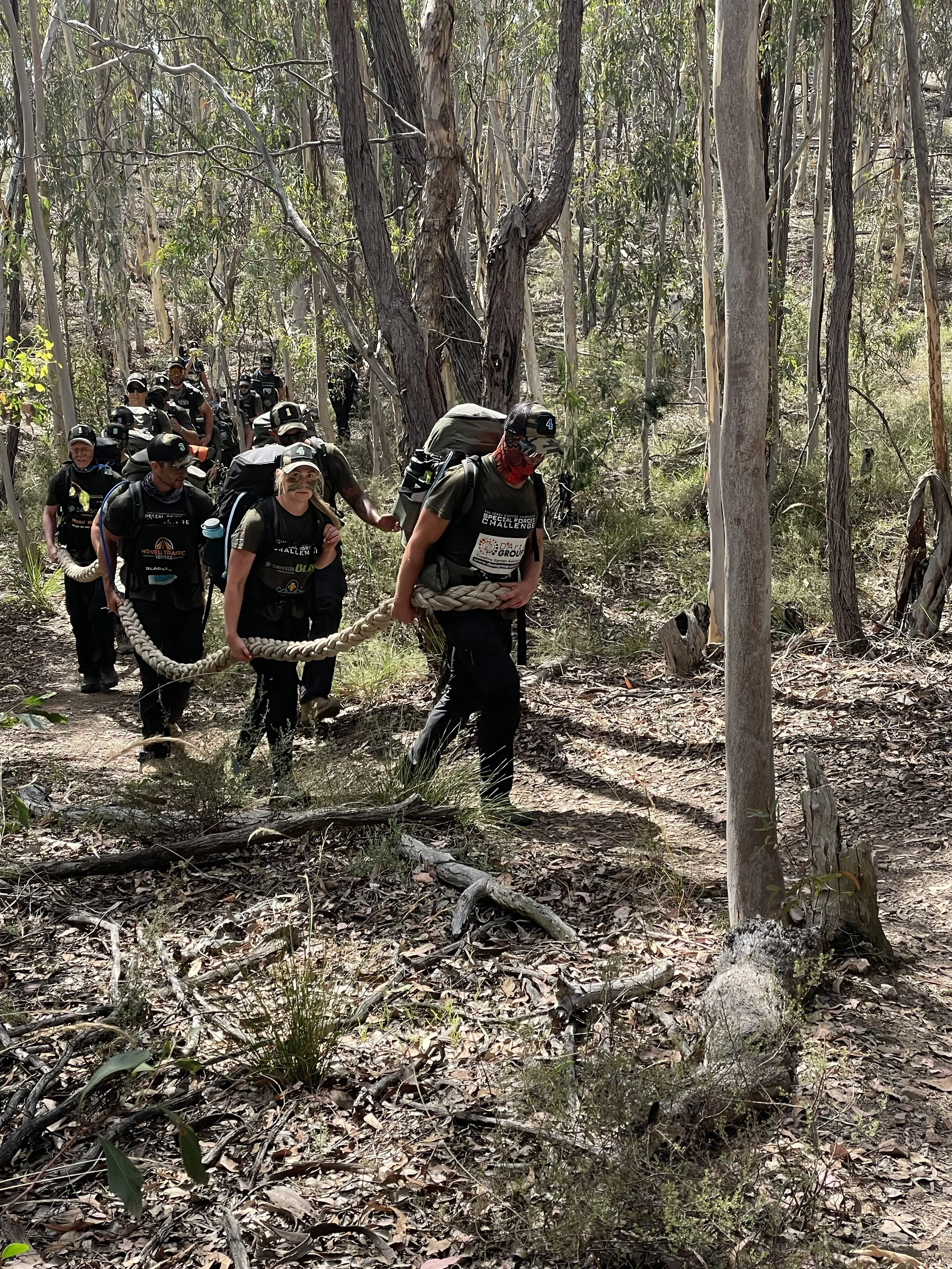 30 Unbroken participants wearing uniforms and backpacks, walking in a forested area with tall trees and dry leaves on the ground.