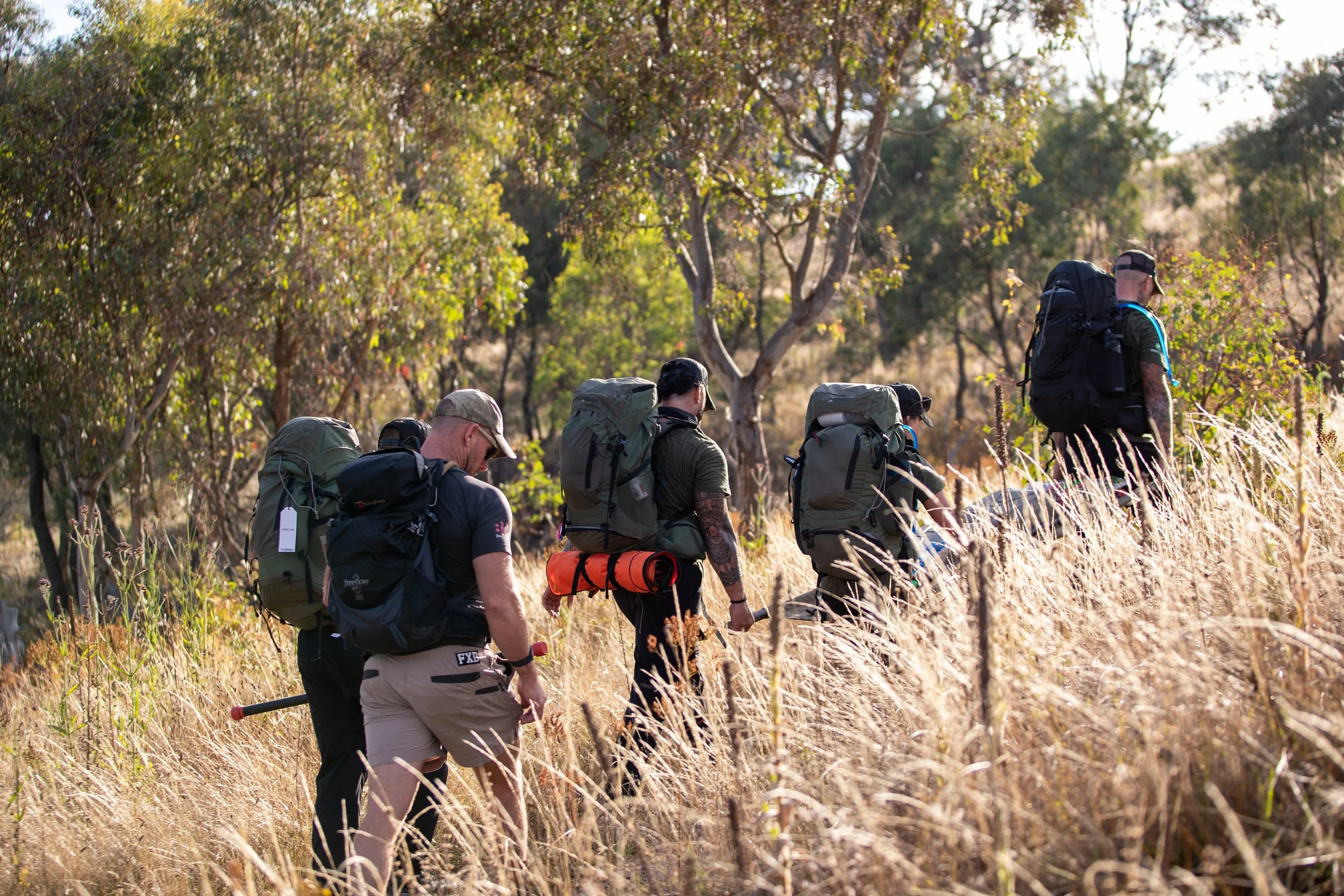 30 Unbroken participants walking through a grassy field in a wooded area during daytime, carrying backpacks.