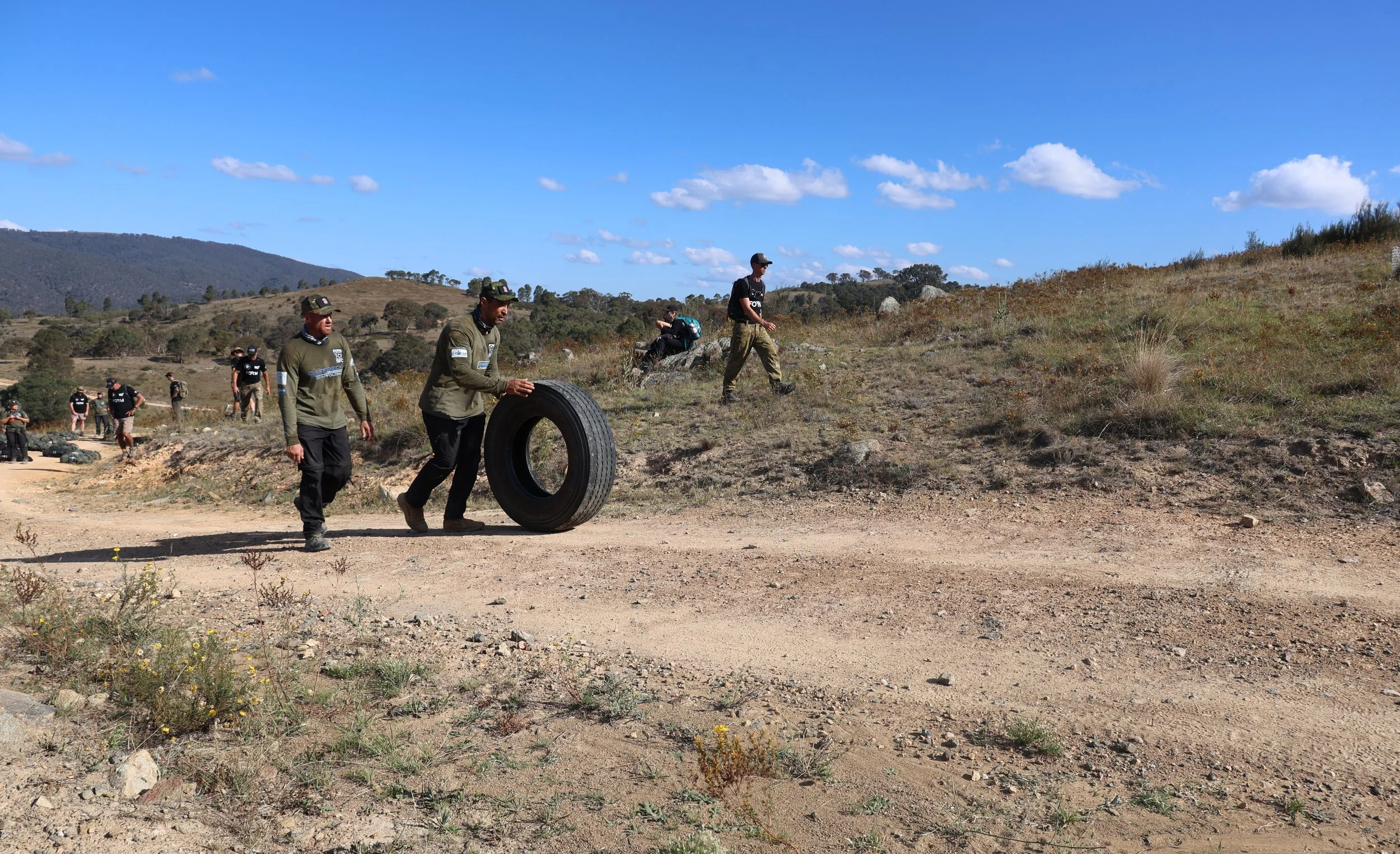 30 Unbroken participants outdoors in a rural, hilly area under a blue sky, with some individuals carrying tires and others walking along the dirt path.