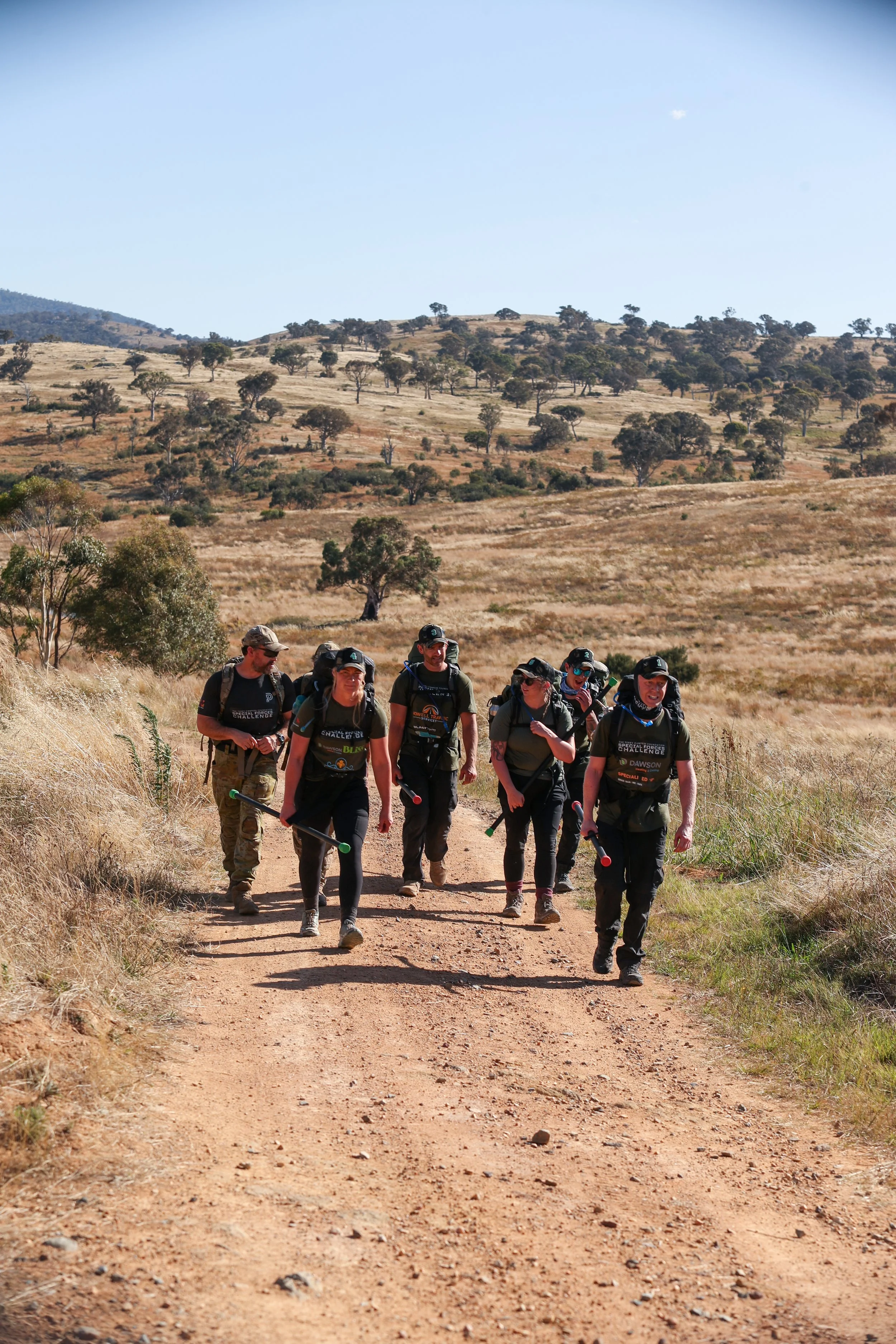 30 Unbroken participants walking on a dirt trail in a hilly, dry landscape with sparse trees under a clear blue sky.