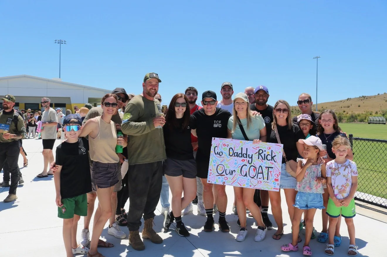Group of people, including children and adults, gathered outdoors on a sunny day holding a colorful sign that reads "To Daddy Rick, Our GOAT." They are smiling and posing together, with some wearing sunglasses and casual summer clothes.