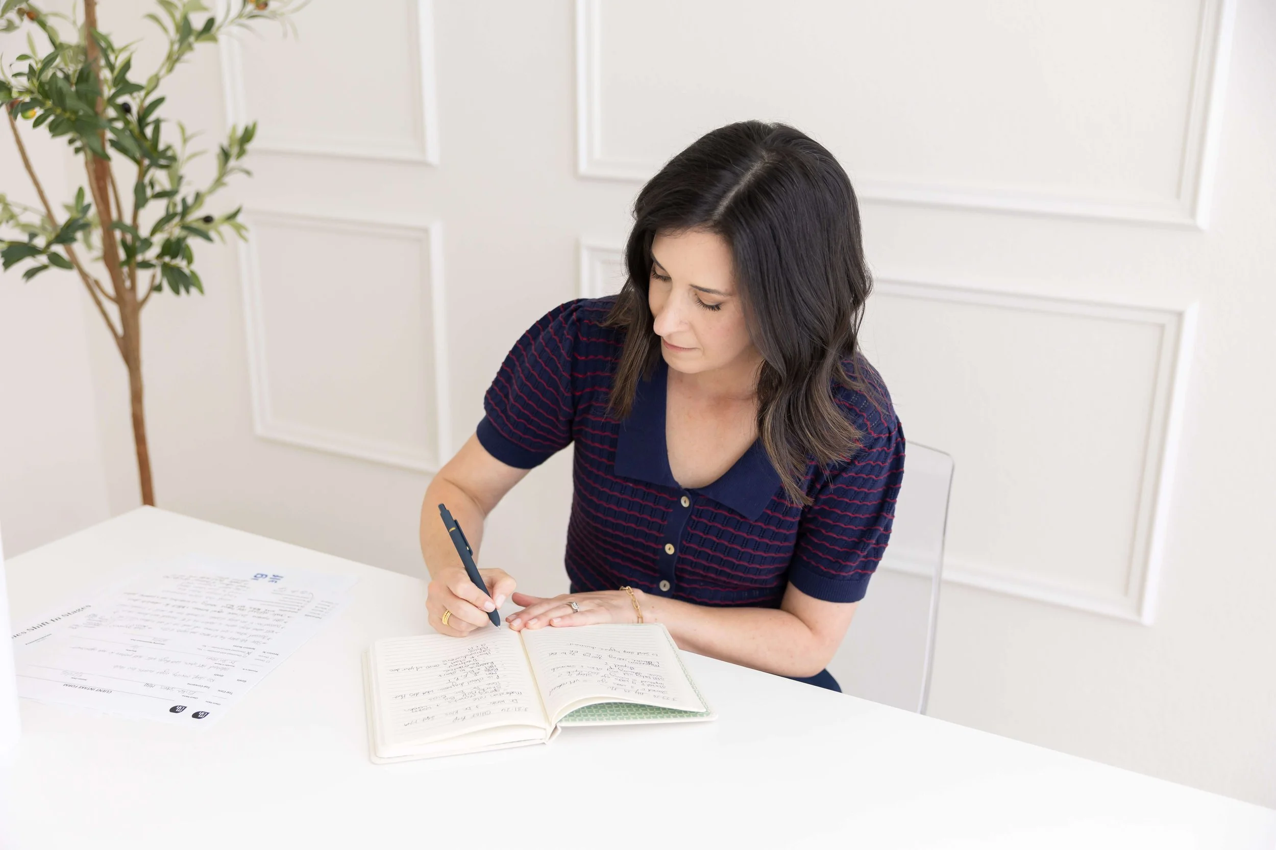 Woman sitting at a white table, writing in a notebook with papers and documents on the table, in a bright room with white walls and a potted plant.