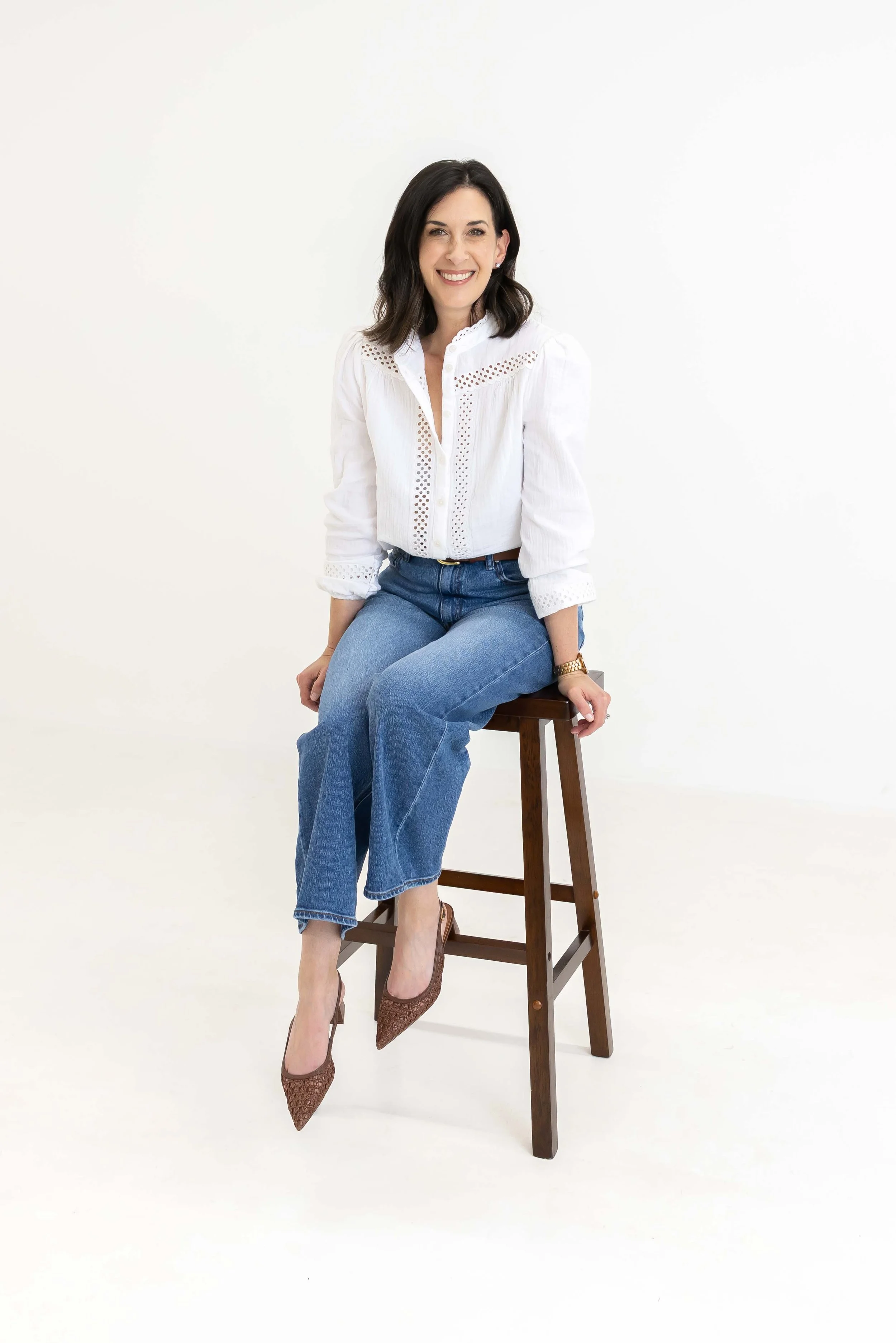 A woman with dark hair is sitting on a wooden stool against a plain white background. She is smiling and dressed in a white button-up shirt with decorative cutouts, blue jeans, and brown high heels. She is wearing a gold watch and earrings.