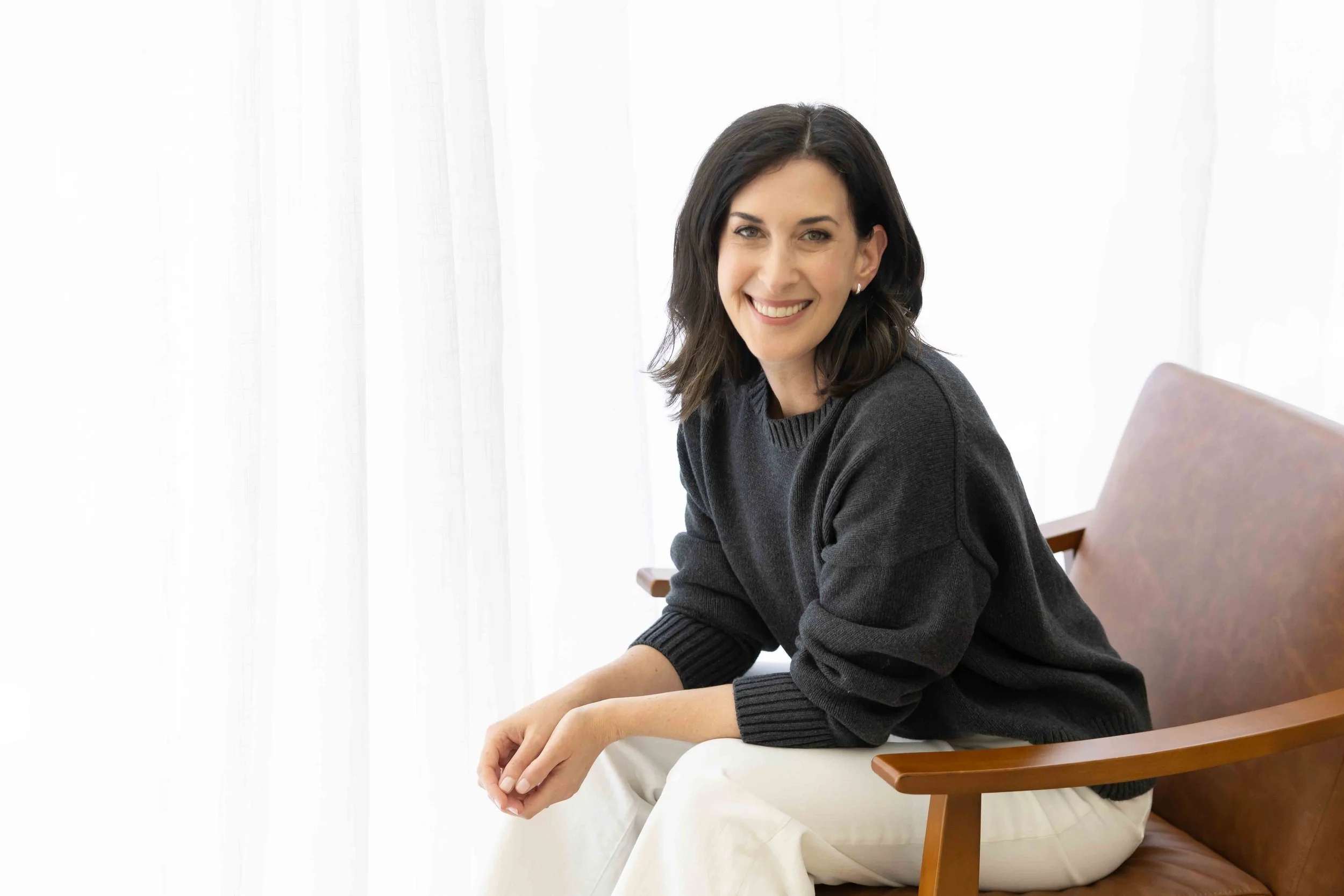 A woman with dark hair and a smile, wearing a dark sweater, seated in a wooden chair with a white curtain background.