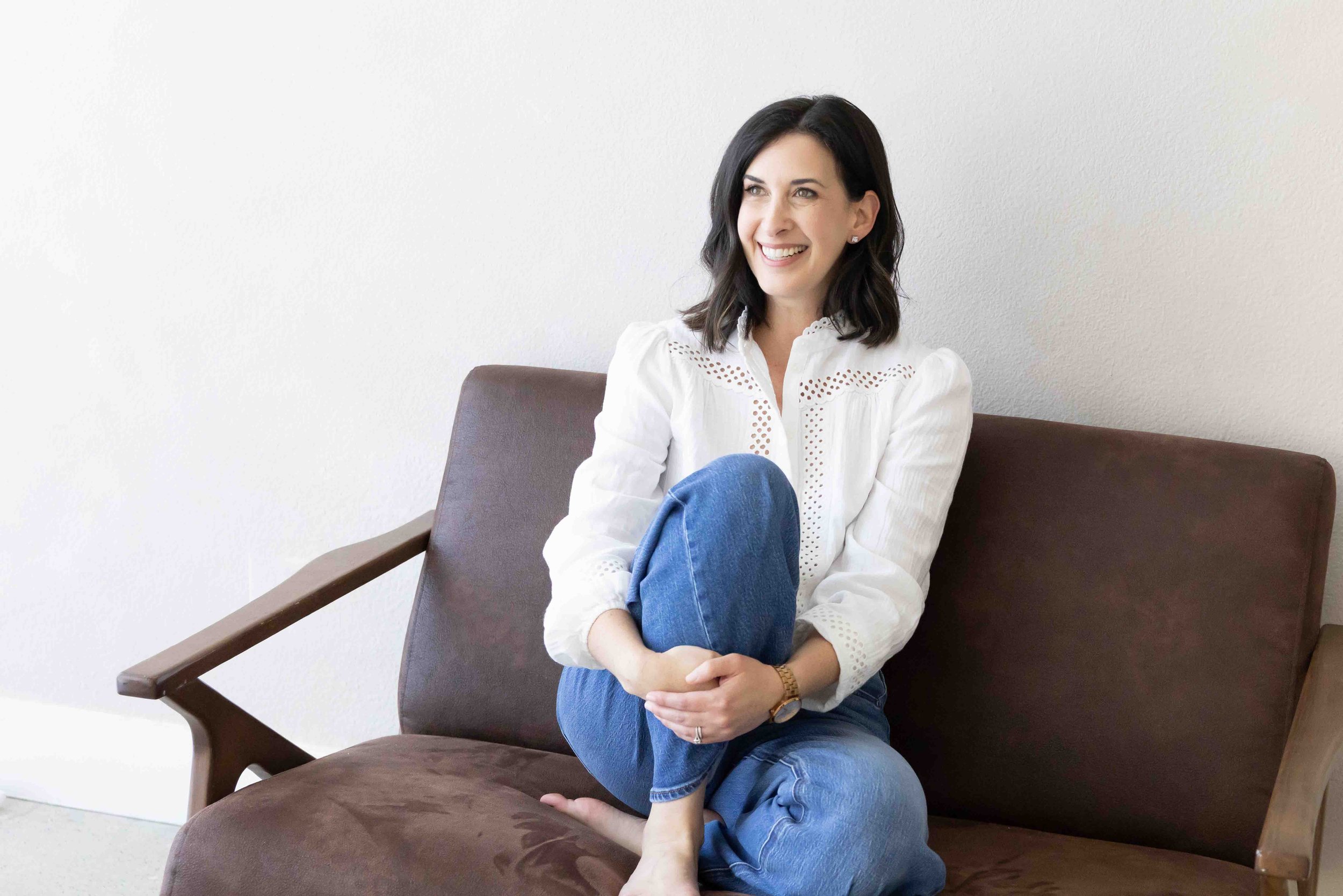 A woman sitting on a brown bench with a white wall in the background. She has black hair, is smiling, and is dressed in a white blouse and blue jeans.